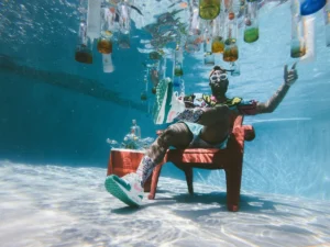 Person sitting underwater in a swimming pool on a red plastic chair, wearing goggles and colorful clothing, surrounded by suspended glass bottles and poolside props in a surreal artistic setup